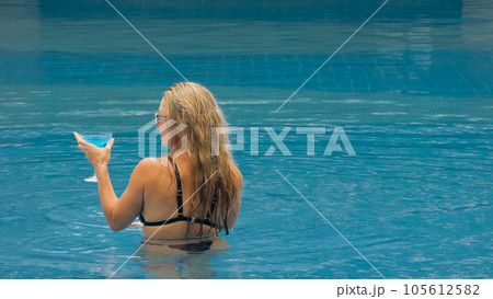 The wide middle shot pretty woman drinking blue cocktail alcohol liquor in swimming pool at hotel. Portrait of sexy girl in pool outdoor. 105612582