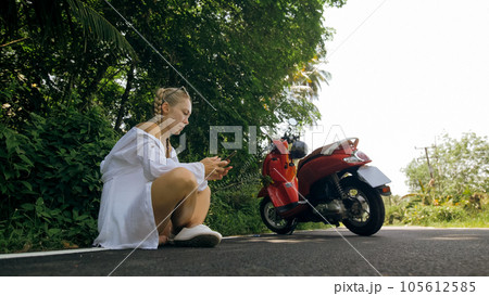 Woman on red motorbike in white clothes drive on forest road trail trip. One girl caucasian tourist sit, near scooter. Use smartphone, take a selfie. Woman on red motorbike in white clothes drive on forest road trail trip. One girl caucasian tourist sit, near scooter. Use smartphone, take a selfie. 105612585