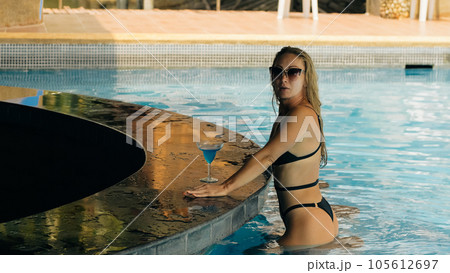 The pretty woman drinking blue cocktail alcohol liquor and sit at the bar. Cocktail bar in the swimming pool. Portrait of sexy girl in pool outdoor. The pretty woman drinking blue cocktail alcohol liquor and sit at the bar. Cocktail bar in the swimming pool. Portrait of sexy girl in pool outdoor. 105612697