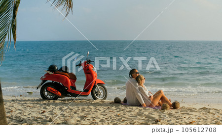 Scooter road trip. Lovely couple on red motorbike in white clothes on sand beach. People walking near the tropical palm trees, sea. Motorcycle rent. Scooter road trip. Lovely couple on red motorbike in white clothes on sand beach. People walking near the tropical palm trees, sea. Motorcycle rent. 105612708