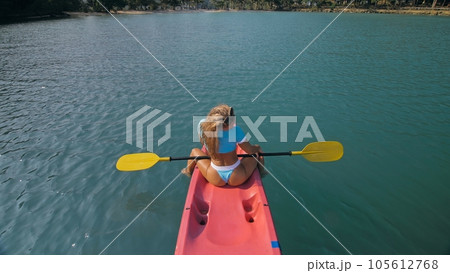Seductive blonde woman in blue swimsuit sails on pink plastic kayak along azure sea at exotic resort backside upper above view. 105612768