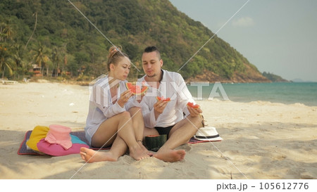 The cheerful love couple holding and eating slices of watermelon on tropical sand beach sea. Romantic lovers two people spend summer weekend. 105612776