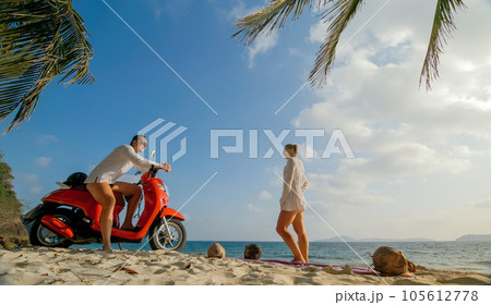 Scooter road trip. Lovely couple on red motorbike in white clothes on sand beach. People walking near the tropical palm trees, sea. Motorcycle rent. 105612778