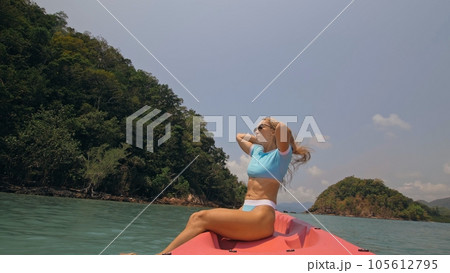 Pretty young blonde woman in stylish swimsuit and sunglasses sits on pink canoe sailing along sea against hills. Pretty young blonde woman in stylish swimsuit and sunglasses sits on pink canoe sailing along sea against hills. 105612795