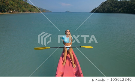 Pretty sportive blonde lady in stylish swimsuit sails on pink plastic canoe along azure ocean bay at exotic resort. 105612840