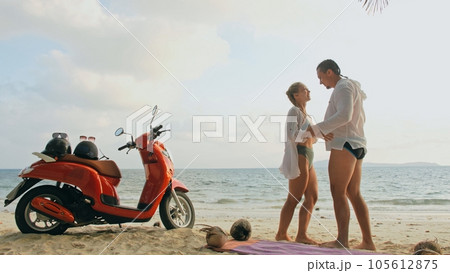 Scooter road trip. Lovely couple on red motorbike in white clothes on sand beach. People walking near the tropical palm trees, sea. Motorcycle rent. Scooter road trip. Lovely couple on red motorbike in white clothes on sand beach. People walking near the tropical palm trees, sea. Motorcycle rent. 105612875