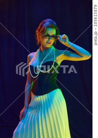 Portrait of woman, young fashion model in fashionable outfit touching sunglasses posing over dark violet background in neon light Portrait of woman, young fashion model in fashionable outfit touching sunglasses posing over dark violet background in neon light 105615879