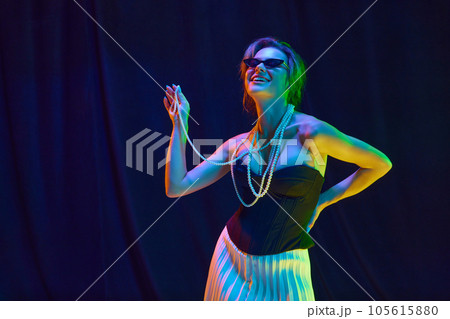 Dreaming and smiling woman, young fashion model in fashionable outfit touching bead necklace posing over dark violet background in neon light Dreaming and smiling woman, young fashion model in fashionable outfit touching bead necklace posing over dark violet background in neon light 105615880