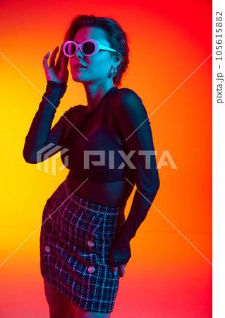 Portrait of woman wearing fashionable touching sunglasses in transparent top and skirt posing over gradient yellow-red neon background. Portrait of woman wearing fashionable touching sunglasses in transparent top and skirt posing over gradient yellow-red neon background. 105615882