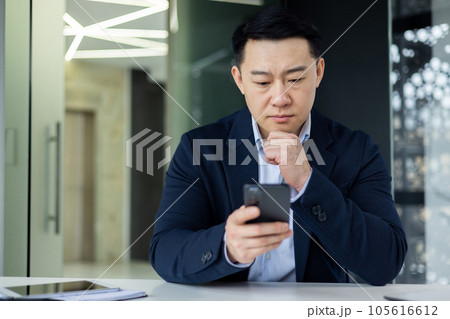 Mature thinking asian boss inside office workplace with phone in hands, successful businessman thinking sitting with laptop in business clothes. Mature thinking asian boss inside office workplace with phone in hands, successful businessman thinking sitting with laptop in business clothes. 105616612