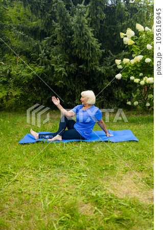 Athletic 50s senior woman doing poses at yoga mat outdoors dressed in sport clothes alone in the park or forest in nature  105616945