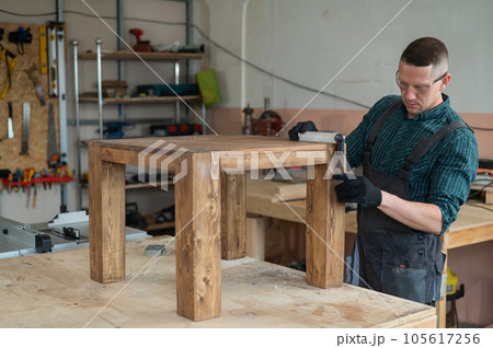 Male carpenter finishing work on wooden table in workshop. Male carpenter finishing work on wooden table in workshop. 105617256