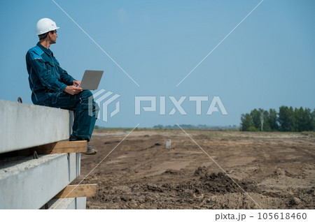 Caucasian male builder in hardhat sits on floor slabs and uses laptop at construction site.  105618460