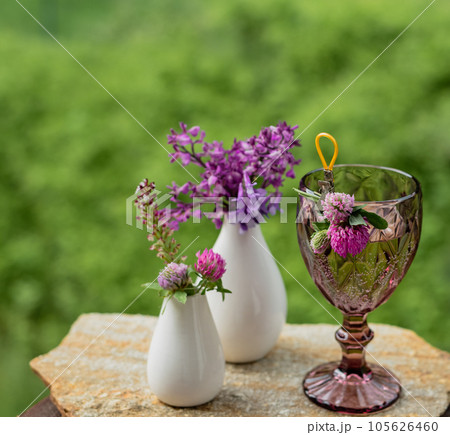 A glass with a drink and flowers in a white vase on the terrace against the backdrop of a green garden A glass with a drink and flowers in a white vase on the terrace against the backdrop of a green garden 105626460