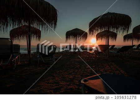 First Rays Of Sun Mark Beginning Of New Day For Resort. In Background, Silhouettes Of Mountains Of Balkan Peninsula, In Foreground, Silhouette Of Beach And Sun Loungers With Umbrellas. 105631278
