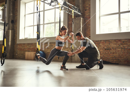 Doing squat exercise. Confident young personal trainer is showing slim athletic woman how to do squats with Trx fitness straps while training at gym. 105631974