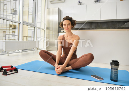 Sport and wellbeing. Young brunette fitness girl, wearing activewear, sitting on yoga mat, meditating, practice relaxation and mindfulness at home, workout in kitchen 105636762