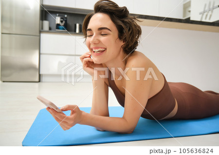 Close up portrait of fitness girl, lying on yoga mat and laughing, using smartphone, records workout exercises on mobile phone, does training at home Close up portrait of fitness girl, lying on yoga mat and laughing, using smartphone, records workout exercises on mobile phone, does training at home 105636824
