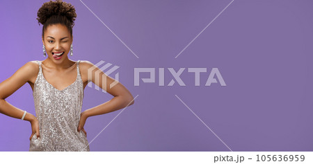 Cheeky good-looking african-american woman in silver glittering dress hold hand waist show tongue daring winking camera feel lucky standing thrilled dancing have fun partying, blue background 105636959