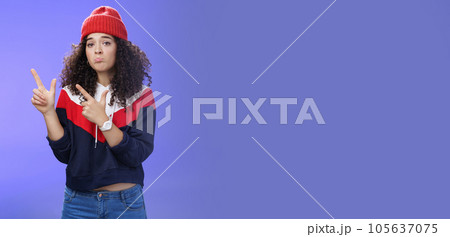 Portrait of silly upset cute curly-haired woman in trendy beanie pursing lips in sadness and frowning making disappointed, regret face pointing at upper left corner unhappy and sad over blue wall Portrait of silly upset cute curly-haired woman in trendy beanie pursing lips in sadness and frowning making disappointed, regret face pointing at upper left corner unhappy and sad over blue wall 105637075