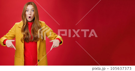 Impressed girl cannot believe what sees open mouth from surprise and excitement popping eyes questioned at camera, pointing down with index fingers asking question and being astonished over red wall 105637373