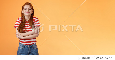 Tender lovely redhead modern woman embrace own body, feel happiness self-acceptance, smiling close eyes, cross arms, hugging herself dreamy, imaging something, daydreaming orange background 105637377