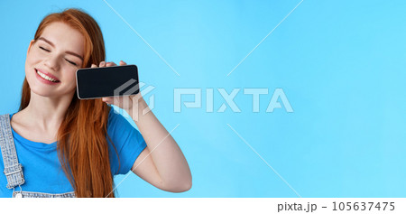 Carefree tender redhead european female tilt head, smiling closed eyes, hold smartphone horizontally, show loving girlfriend photos, romantic video online, stand blue background cheerful 105637475