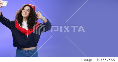 Cute girl making selfie to memorize trip standing in warm winter beanie and sweatshirt posing as pulling hand with smartphone and smiling at device screen to take pretty shot and post internet 105637535