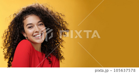 Romantic and flirty young cute caucasian girlfriend with curly hair in red blouse making silly pose smiling joyfully and lifting shoulder coquettish smiling broadly at camera over yellow wall 105637578