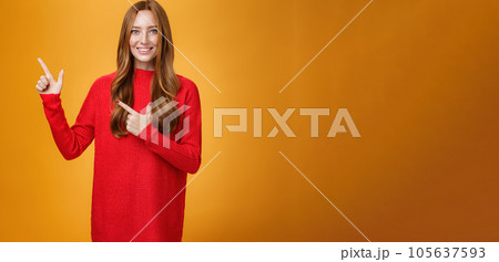 Assertive and helpful charming young female assitant in red knitted dress with ginger hair smiling friendly and pleasant as pointing at upper left corner, showing interesting thing over orange wall Assertive and helpful charming young female assitant in red knitted dress with ginger hair smiling friendly and pleasant as pointing at upper left corner, showing interesting thing over orange wall 105637593