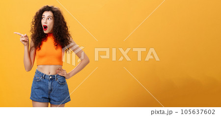 Woman peeking at neighbours window being surprised and amazed pointing and staring speechless left standing over orange background with dropped jaw intrigued and thrilled look in cropped top 105637602