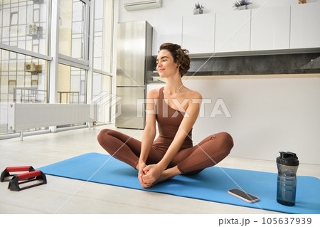 Portrait of fitness girl doing yoga on rubber mat at home, workout indoors in kitchen wearing sportswear, practice minfulness, sitting in lotus pose with water bottle next to her 105637939