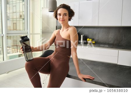 Portrait of smiling fitness girl, wearing sportswear for jogging, workout training, standing in kitchen and drinking water from sports bottle, looking fit and happy Portrait of smiling fitness girl, wearing sportswear for jogging, workout training, standing in kitchen and drinking water from sports bottle, looking fit and happy 105638010