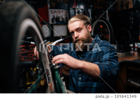 Closeup portrait of bearded repairman fixing mountain bicycle standing on bike rack working in bike repair shop with dark interior, looking at camera. Concept of maintenance of bicycle transport. 105641349