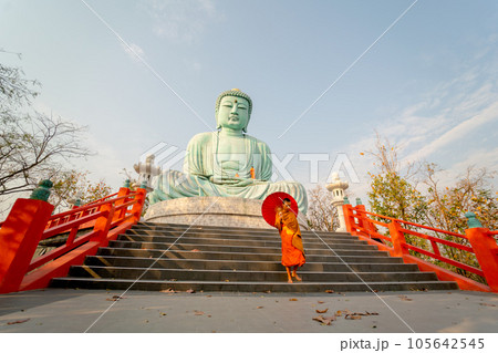 Wide shot and side view of young Asian monk hold red umbrella and walk down on stair in front statue 105642545