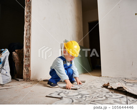 Little boy construction worker installing ceramic tile on the floor in apartment under renovation. Kid wearing safety helmet and work overalls while playing at home. Little boy construction worker installing ceramic tile on the floor in apartment under renovation. Kid wearing safety helmet and work overalls while playing at home. 105644446
