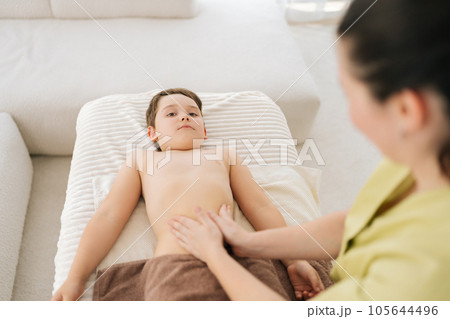 High-angle view of five year old boy patient having therapeutic massage on tummy lying on couch in medical clinic. Closeup hands of unrecognizable female pediatric masseuse massaging tummy of child. 105644496