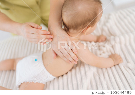 Closeup top view of loving mother hands massaging little baby. Close up cropped shot of unrecognizable masseuse female hands doing massage to newborn. Back view of cute infant baby lying on stomach. 105644499
