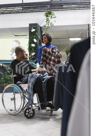 Clothing store asian woman customer showing plaid shirt on hanger to arab man with physical impairment. Mall boutique client in wheelchair getting assistance from seller while shopping for apparel Clothing store asian woman customer showing plaid shirt on hanger to arab man with physical impairment. Mall boutique client in wheelchair getting assistance from seller while shopping for apparel 105646867