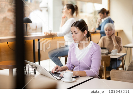 Positive European young woman drinking and enjoying coffee while working on laptop in cafe. Freelance and remote work 105648311