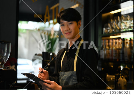 Young male waitress wearing apron standing behind counter and taking order from customer in cafe or restaurant 105660022