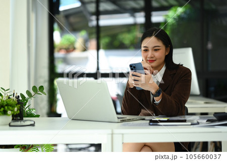 Beautiful young businesswoman sitting in front of laptop at office desk and communicating online on mobile phone 105660037