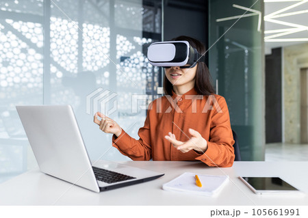 Asian woman wearing virtual reality glasses sitting at a desk in a workspace inside the office, businesswoman in online meeting talking remotely with colleagues, virtual meeting simulation. 105661991