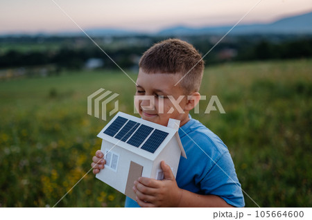 Boy with model house with installed of solar panels, standing in the middle of meadow. 105664600