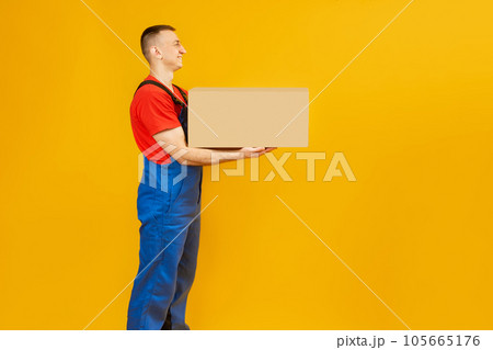 Man in blue jumpsuit holds cardboard box on yellow studio background. Goods delivery courier. Side view. Copy space, mock up. Man in blue jumpsuit holds cardboard box on yellow studio background. Goods delivery courier. Side view. Copy space, mock up. 105665176