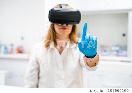Blond haired chemist in VR glasses moves finger imitating touch. Curious blonde woman in rubber gloves tries virtual reality in modern laboratory Blond haired chemist in VR glasses moves finger imitating touch. Curious blonde woman in rubber gloves tries virtual reality in modern laboratory 105666160