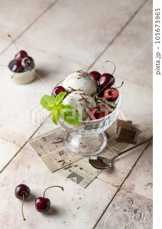 Ice cream in balls with cherry, chocolate and mint in glass bowl on beige tile background, summer refreshing morning dessert, vertical 105673965