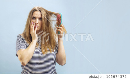 Very sad and upset young woman looking with shock at her damaged hair on blue background 105674373