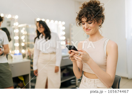 Smiling young woman with perfect makeup and hairstyle typing online message using smartphone looking to screen, on blurred background of cheerful makeup artist and hairdresser in modern beauty studio. Smiling young woman with perfect makeup and hairstyle typing online message using smartphone looking to screen, on blurred background of cheerful makeup artist and hairdresser in modern beauty studio. 105674467