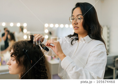 Side view of hairstylist making waves curling on woman hair. Closeup of female hairstylist in beauty salon takes lock of hair and presses with hair straightener. Concept of fashion and beauty. Side view of hairstylist making waves curling on woman hair. Closeup of female hairstylist in beauty salon takes lock of hair and presses with hair straightener. Concept of fashion and beauty. 105674468